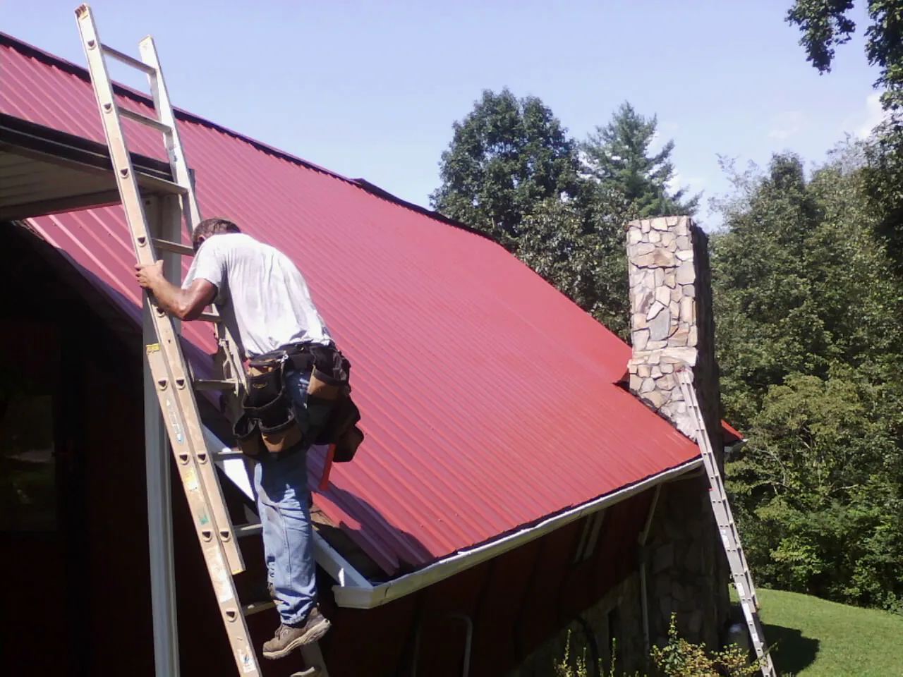 Roofer on a ladder beside a red metal roof and stone chimney — Soffit & Fascia Repair by Roofing Brothers in Parrottsville